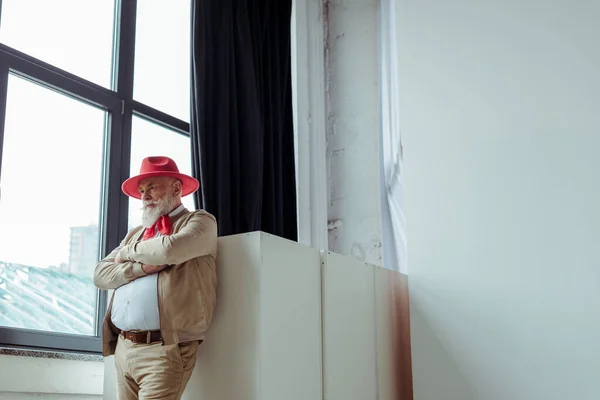 Elegante hombre mayor en sombrero rojo de pie cerca de la ventana en el estudio de fotos - foto de stock