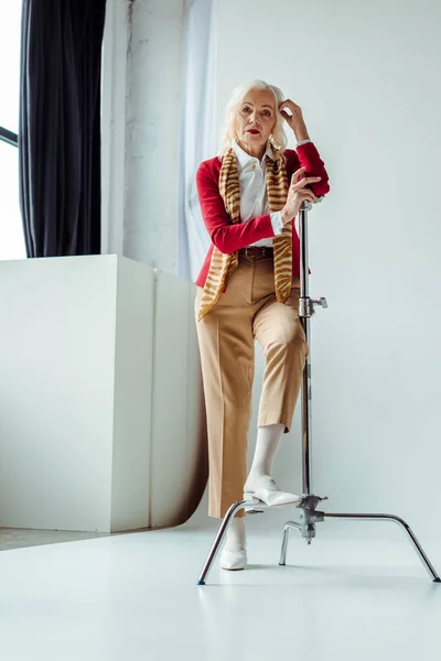 Elegant senior woman standing near tripod on white background in photo studio — Stock Photo