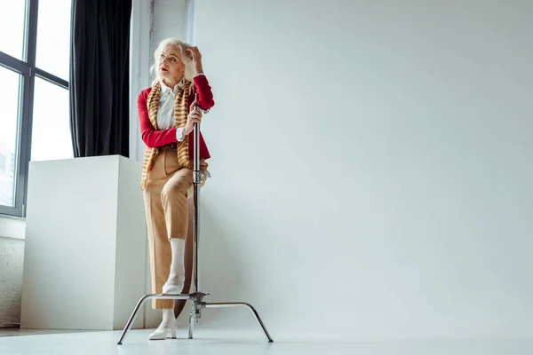 Mulher idosa bonita olhando para longe perto de tripé no fundo branco no estúdio de fotos — Fotografia de Stock