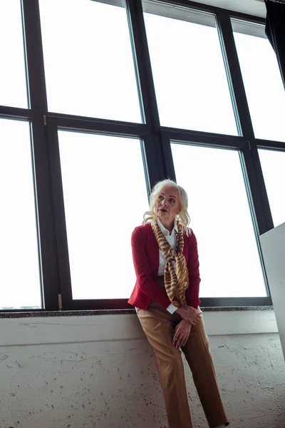 Elegante mujer mayor mirando hacia otro lado cerca de la ventana en el estudio de fotos - foto de stock