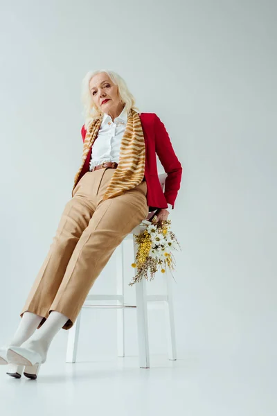 Mulher idosa bonita com flores silvestres olhando para a câmera na cadeira no fundo branco — Fotografia de Stock