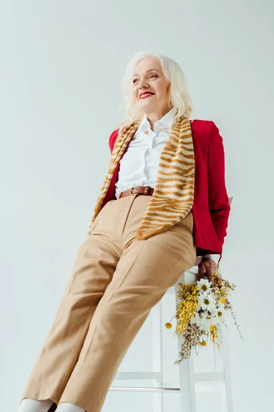 Elegante mujer mayor sosteniendo flores silvestres y sonriendo en silla aislada en blanco - foto de stock