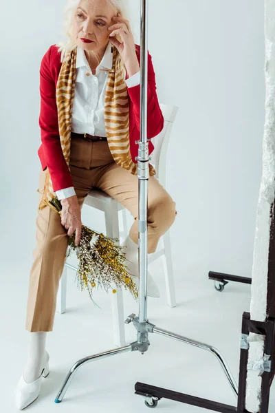 Mulher sênior elegante com flores silvestres sentado na cadeira perto de tripé no fundo branco — Fotografia de Stock