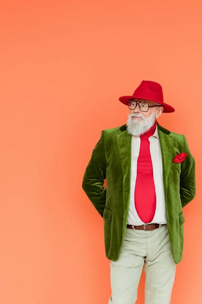 Fashionable elderly man in hat and eyeglasses looking away isolated on coral — Stock Photo