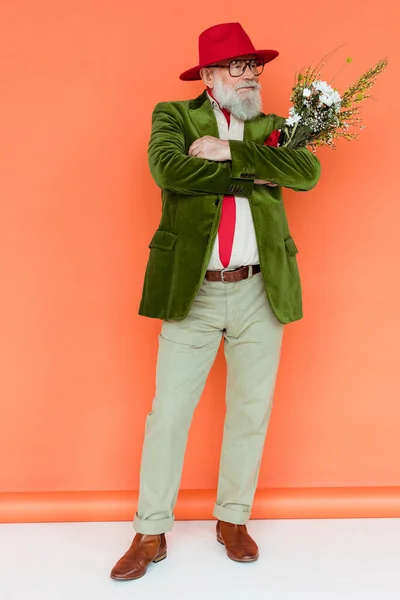 Full length of handsome elderly man with crossed arms holding wildflowers on white surface on coral background — Stock Photo