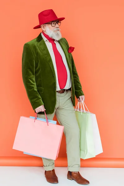 Volle Länge des eleganten älteren Mannes mit Einkaufstaschen auf weißer Oberfläche auf korallenfarbenem Hintergrund — Stockfoto