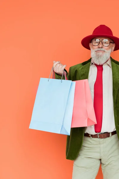 Handsome senior man in hat and eyeglasses holding shopping bags and looking at camera isolated on coral — Stock Photo