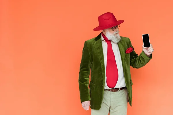 Hombre mayor guapo en sombrero y gafas con teléfono inteligente con pantalla en blanco aislado en coral - foto de stock