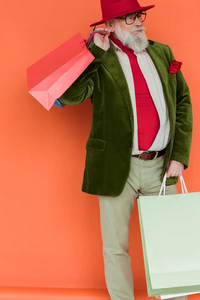 Fashionable elderly man holding shopping bags and looking away on coral background — Stock Photo