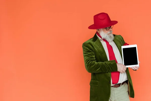 Handsome elderly man in hat and eyeglasses holding digital tablet isolated on coral with copy space — Stock Photo