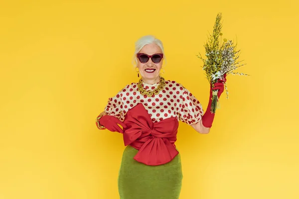 Elegante mujer mayor sonriendo mientras sostiene ramo de flores silvestres aisladas en amarillo - foto de stock