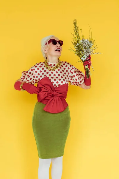 Mujer anciana de moda sosteniendo flores silvestres aisladas en amarillo - foto de stock