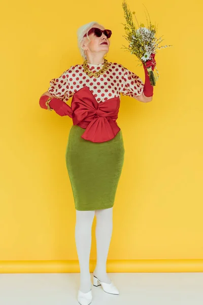 Mulher sênior na moda em óculos de sol que mantêm buquê de flores silvestres na superfície branca no fundo amarelo — Fotografia de Stock
