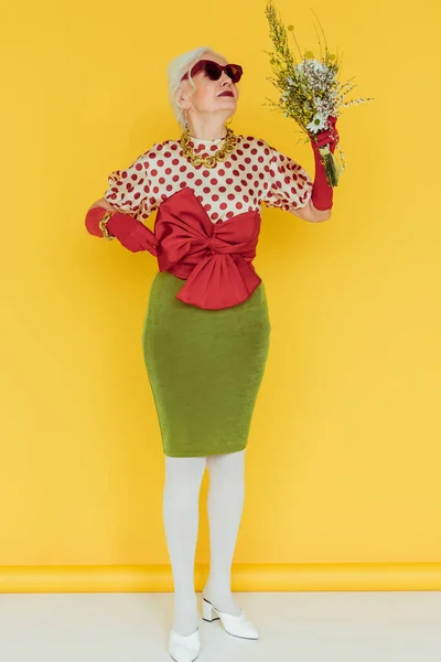 Mujer mayor de moda en gafas de sol con ramo de flores silvestres en la superficie blanca sobre fondo amarillo - foto de stock