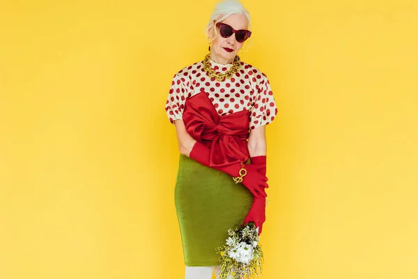 Hermosa anciana en gafas de sol con ramo de flores silvestres aisladas en amarillo - foto de stock