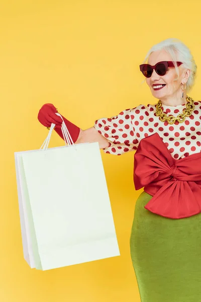 Stylish elderly woman smiling while holding shopping bags isolated on yellow — Stock Photo