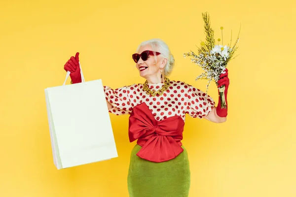 Une dame âgée en vogue qui sourit en tenant des fleurs sauvages et des sacs de magasinage sur fond jaune — Photo de stock