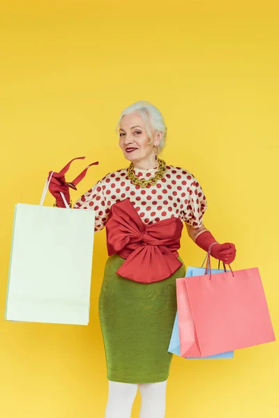 Elegante mujer mayor sonriendo a la cámara mientras sostiene bolsas de compras y gafas de sol aisladas en amarillo - foto de stock