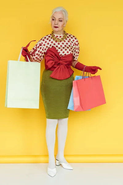 Comprimento total da mulher sênior elegante com sacos de compras e óculos de sol olhando para longe na superfície branca no fundo amarelo — Fotografia de Stock