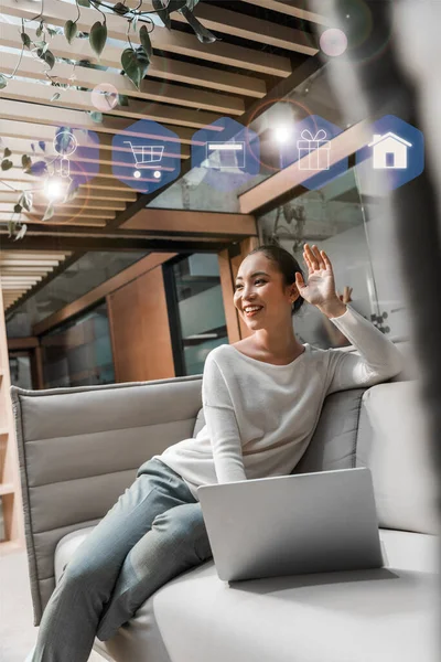 Foyer sélectif de heureux asiatique femme d'affaires agitant la main tout en étant assis sur le canapé près d'un ordinateur portable avec des icônes illustration — Photo de stock