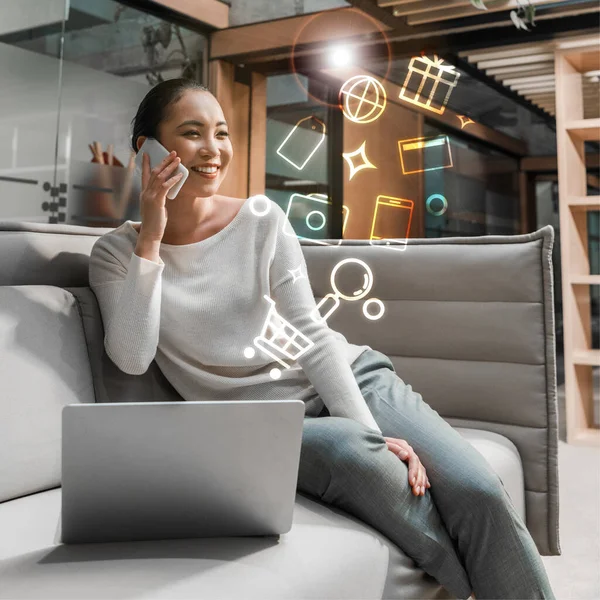 Весела азіатська бізнес-леді розмовляє на смартфоні сидячи на дивані біля ноутбука, ілюстрація цифрових іконок — стокове фото