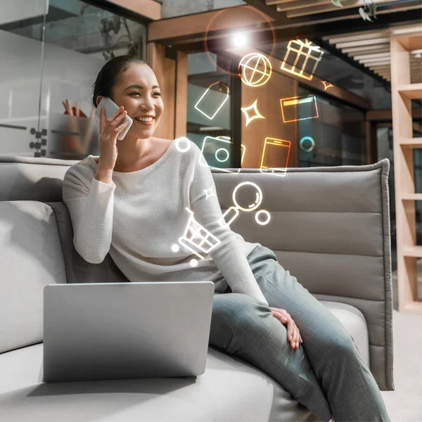 Cheerful asian businesswoman talking on smartphone while sitting on couch near laptop, digital icons illustration — Stock Photo