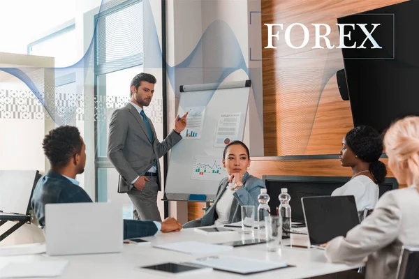 Handsome businessman pointing with finger at flipchart with infographics while multicultural colleagues sitting at desk in conference hall, forex illustration — Stock Photo