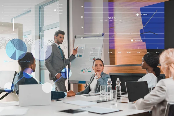 Handsome businessman pointing with finger at flipchart with infographics while multicultural colleagues sitting at desk in conference hall, charts and graphs illustration — Stock Photo