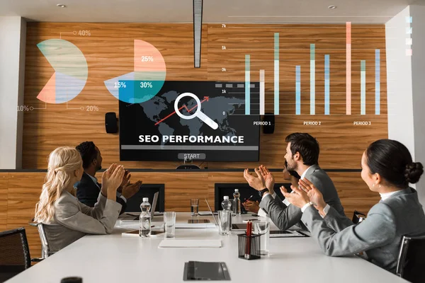 Young multicultural businesspeople applauding while sitting in conference hall and looking at lcd screen on wall, seo performance illustration — Stock Photo