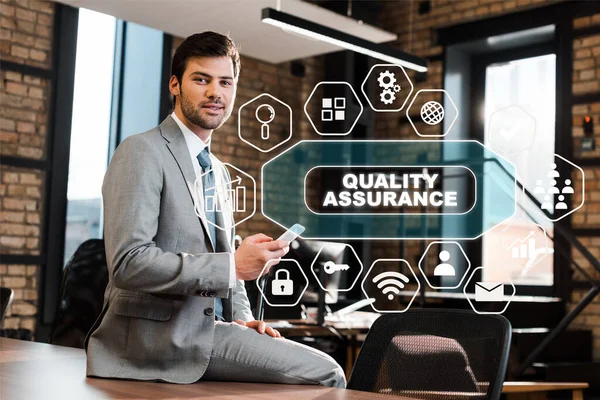 Handsome, positive businessman sitting on office desk, holding smartphone and looking at camera, quality assurance illustration — Stock Photo