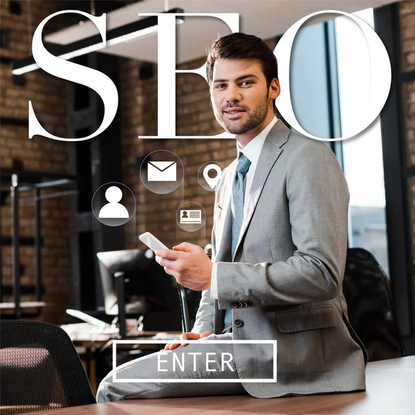 Handsome, positive businessman sitting on office desk, holding smartphone and looking at camera, seo illustration — Stock Photo