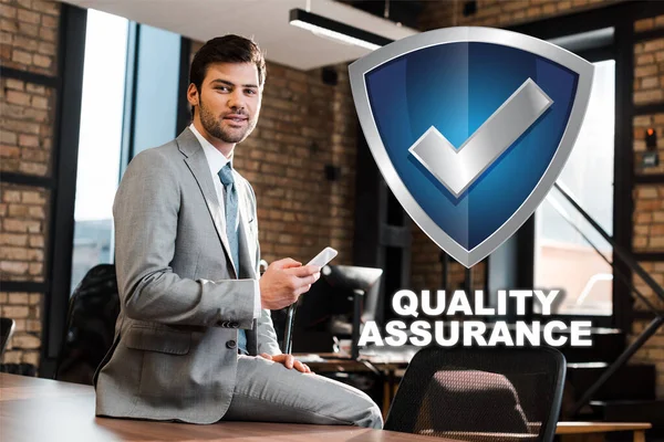 Handsome, positive businessman sitting on office desk, holding smartphone and looking at camera, quality assurance illustration — Stock Photo