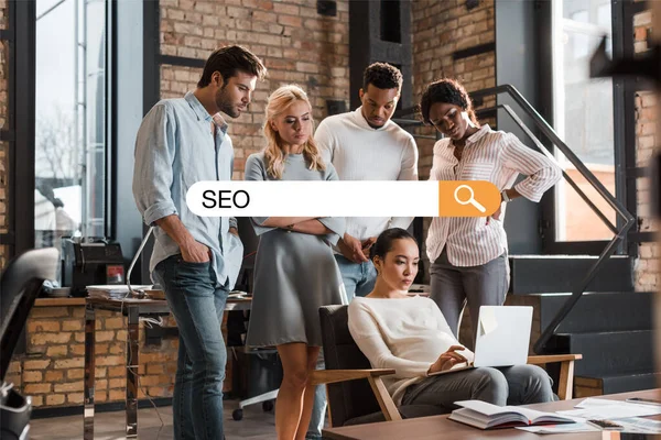 Pensativo multicultural empresarios mirando asiático colega trabajando en portátil, seo ilustración — Stock Photo