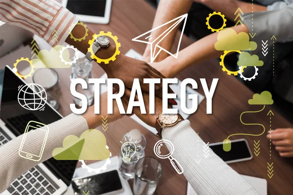 Partial view of multicultural businesspeople holding joined hands above desk with gadgets, strategy illustration — Stock Photo