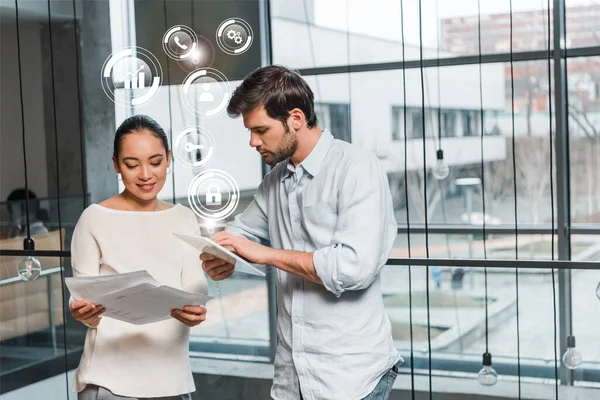 Junger gutaussehender Geschäftsmann mit digitalem Tablet, der neben attraktiven asiatischen Kollegen steht und Dokumente, digitale Symbole und Illustrationen hält — Stockfoto