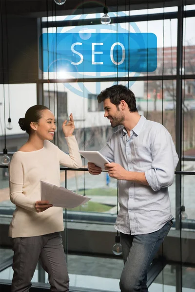 Fröhliche asiatische Geschäftsfrau hält Papiere in der Hand und spricht mit hübschen Kollegen mit digitalem Tablet, seo Illustration — Stockfoto