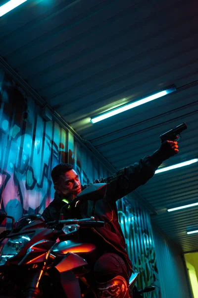 Low angle view of mixed race cyberpunk player on motorcycle aiming gun — Stock Photo