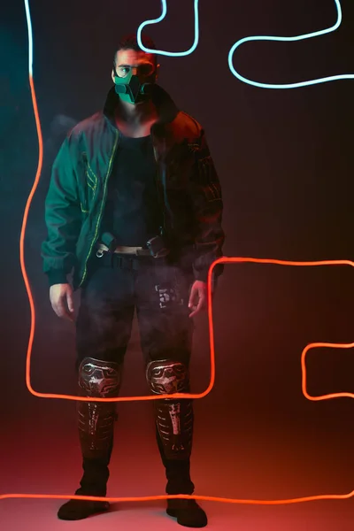 Jugador cyberpunk de raza mixta en máscara protectora de pie cerca de la iluminación de neón rojo en negro - foto de stock