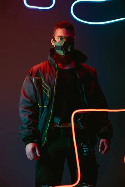 Armed and bi-racial cyberpunk player in protective mask standing near neon lighting on black — Stock Photo