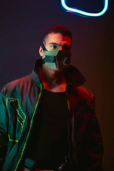 Bi-racial cyberpunk player in protective mask near neon lighting on black — Stock Photo