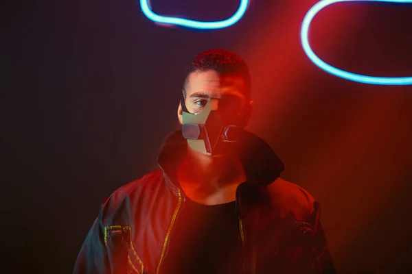 Bi-racial cyberpunk player in protective mask looking at camera near neon lighting on black — Stock Photo