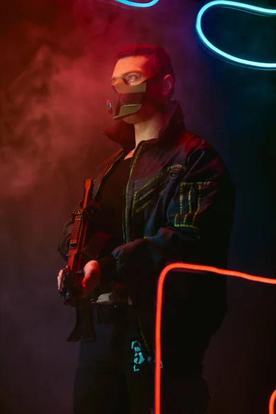 Bi-racial cyberpunk player in protective mask holding gun near neon lighting on black with smoke — Stock Photo