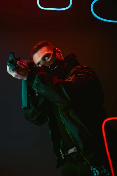 Bi-racial cyberpunk player in protective mask aiming gun near neon lighting on black — Stock Photo