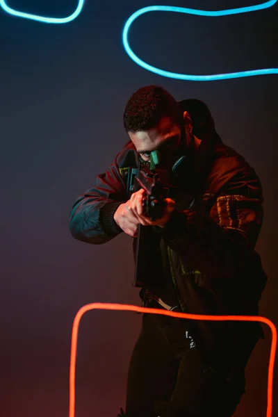 Mixed race cyberpunk player in protective mask aiming gun near neon lighting on black — Stock Photo