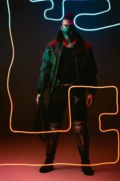 Mixed race cyberpunk player in protective mask holding gun while standing near neon lighting on black — Stock Photo