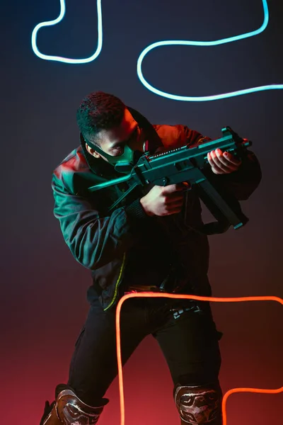 Bi-racial cyberpunk man in protective mask aiming gun near neon lighting on black — Stock Photo