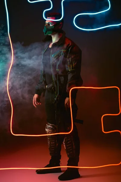 Armed mixed race cyberpunk player in protective mask standing near neon lighting on black with smoke — Stock Photo