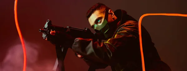 Tiro panorámico de jugador bi-racial cyberpunk en máscara protectora apuntando pistola cerca de la iluminación de neón en negro - foto de stock