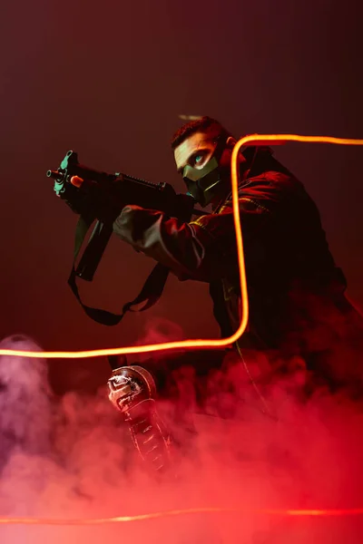 Pericoloso giocatore cyberpunk bi-razziale in maschera protettiva mirando pistola vicino illuminazione al neon su nero con fumo — Foto stock