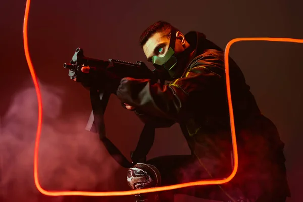 Selective focus of dangerous mixed race cyberpunk player in protective mask aiming gun near neon lighting on black with smoke — Stock Photo